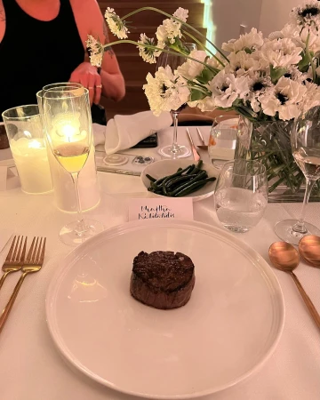 Overhead shot of a plated meat dish on a white tablecloth, complemented by silverware, glassware, candles, flowers, and greenery.