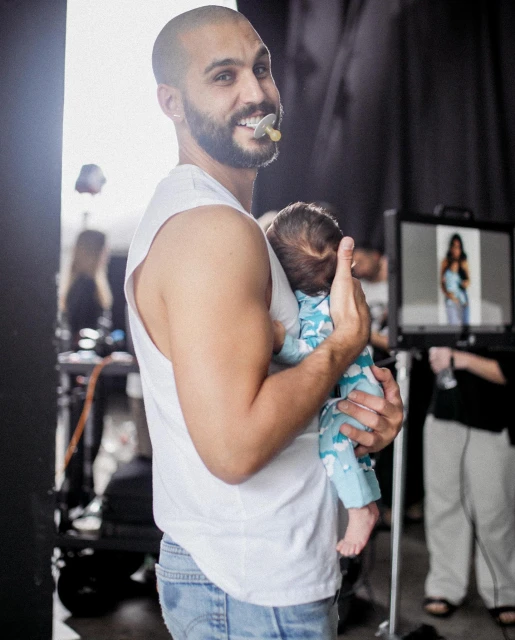 A bald man with a beard is holding a baby in an indoor environment, with studio equipment and lights visible in the background.