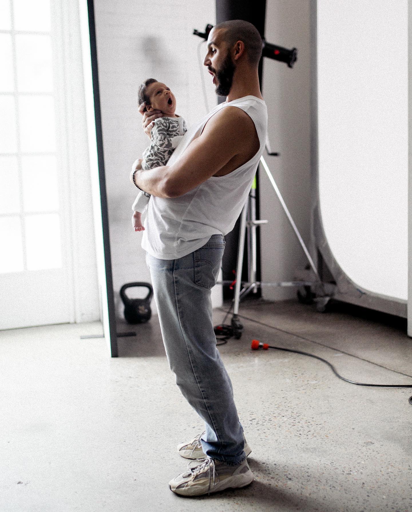 Father holding a yawning baby in photo studio with lighting equipment and white background for a family portrait or lifestyle shoot.