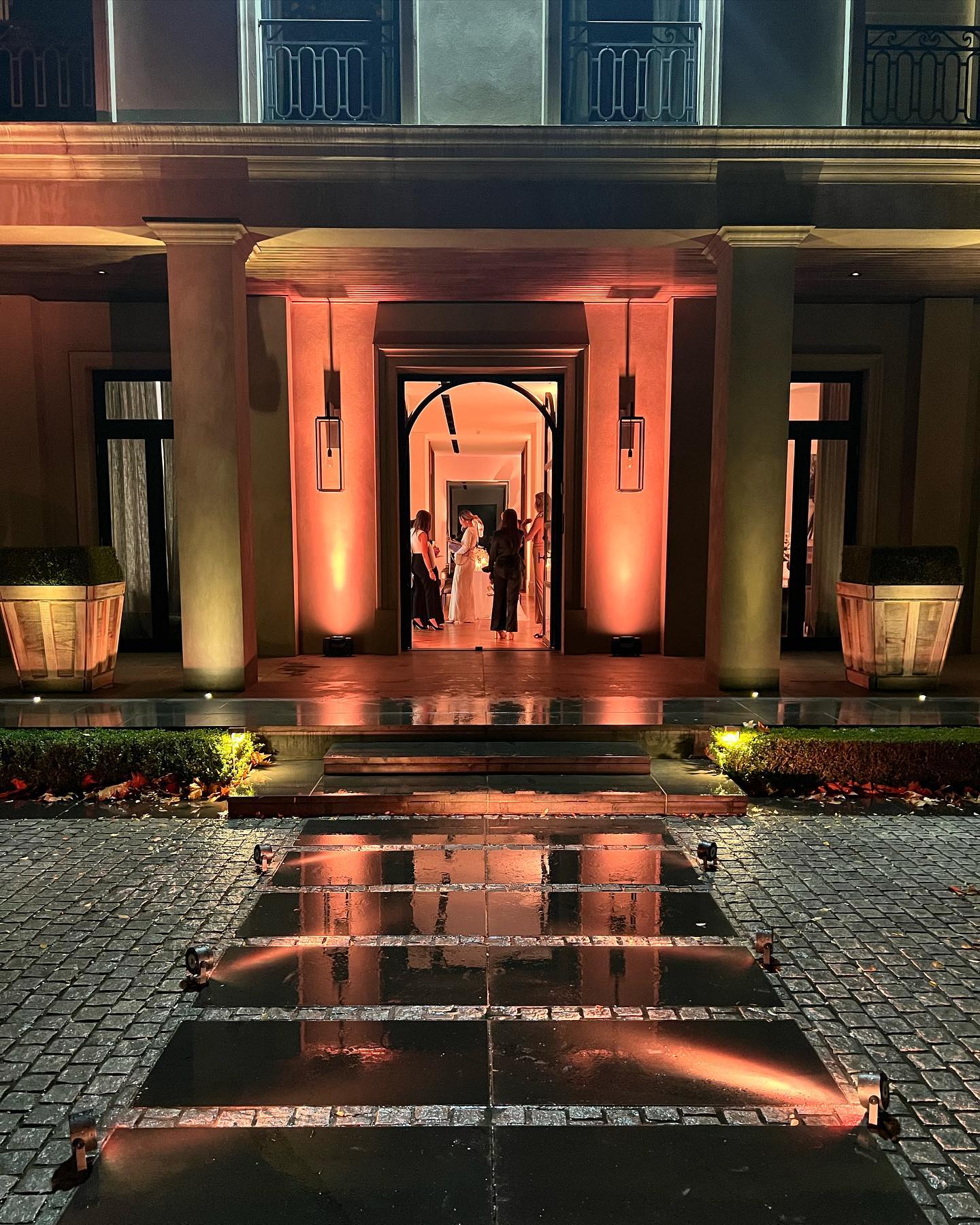 Exterior view of a building facade with an arched entrance, columns, dark pavement, and several silhouetted people standing inside.