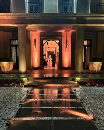 Exterior view of a building facade with an arched entrance, columns, dark pavement, and several silhouetted people standing inside.