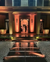 Exterior view of an illuminated building at night with a cobblestone entrance path and people gathered inside an arched doorway.