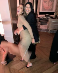 Two women laughing and hugging in a living room, one wearing a beige dress and the other a black blazer.