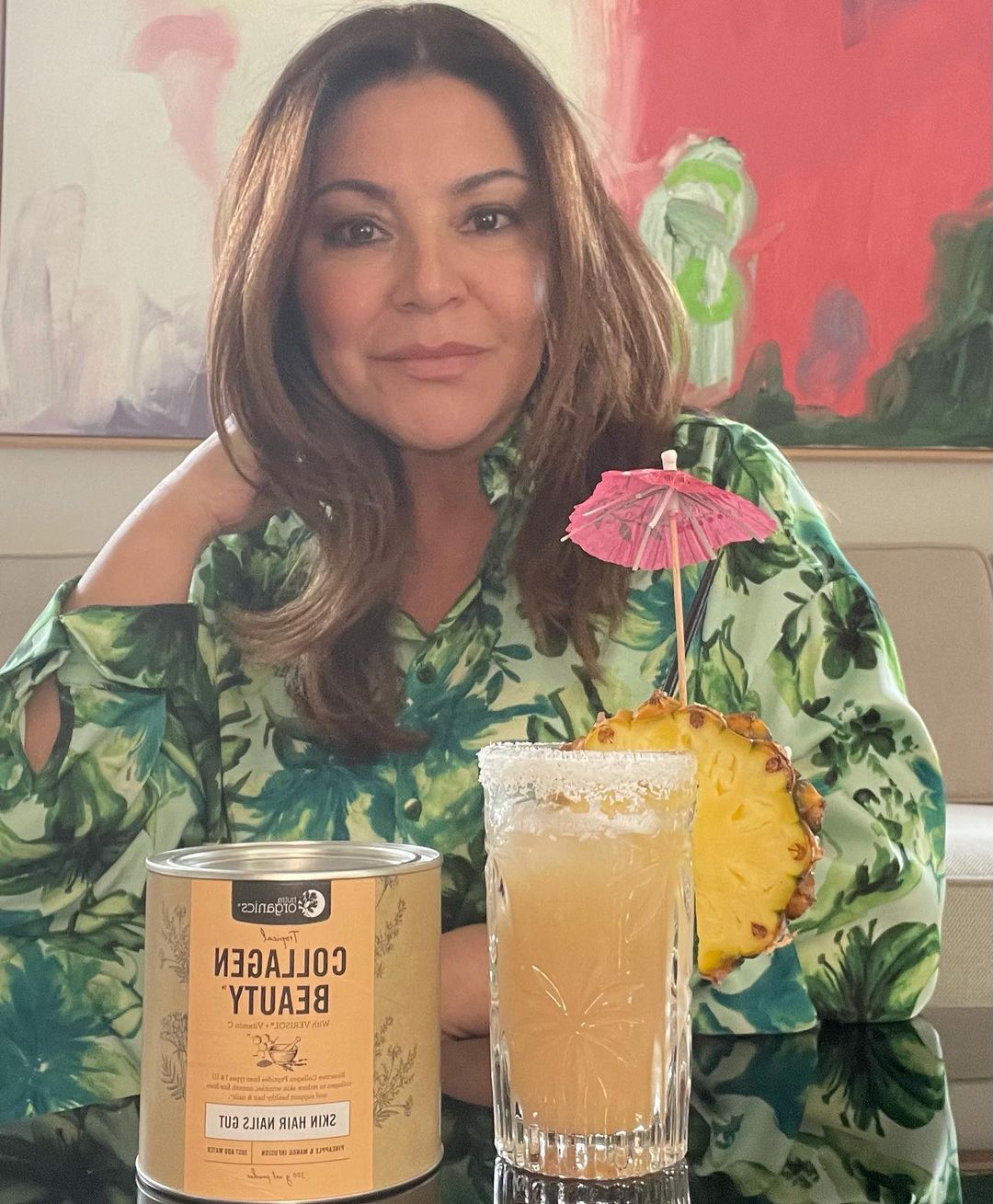 Brunette woman poses indoors with a cocktail, pineapple slice, and collagen beauty supplement on a reflective black table surface.