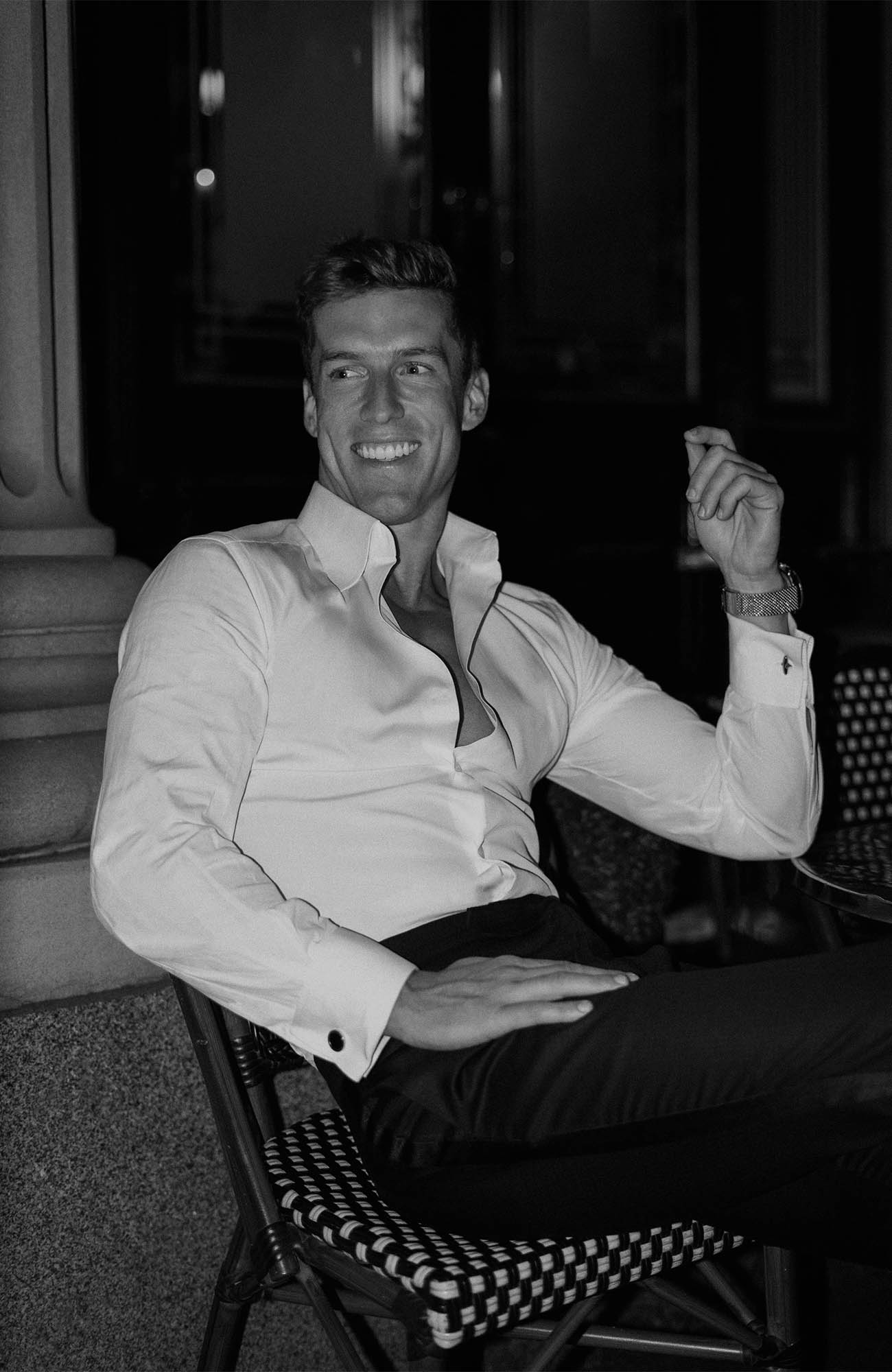 Black and white portrait of an adult male seated on a wicker chair in an outdoor setting, wearing an unbuttoned dress shirt.
