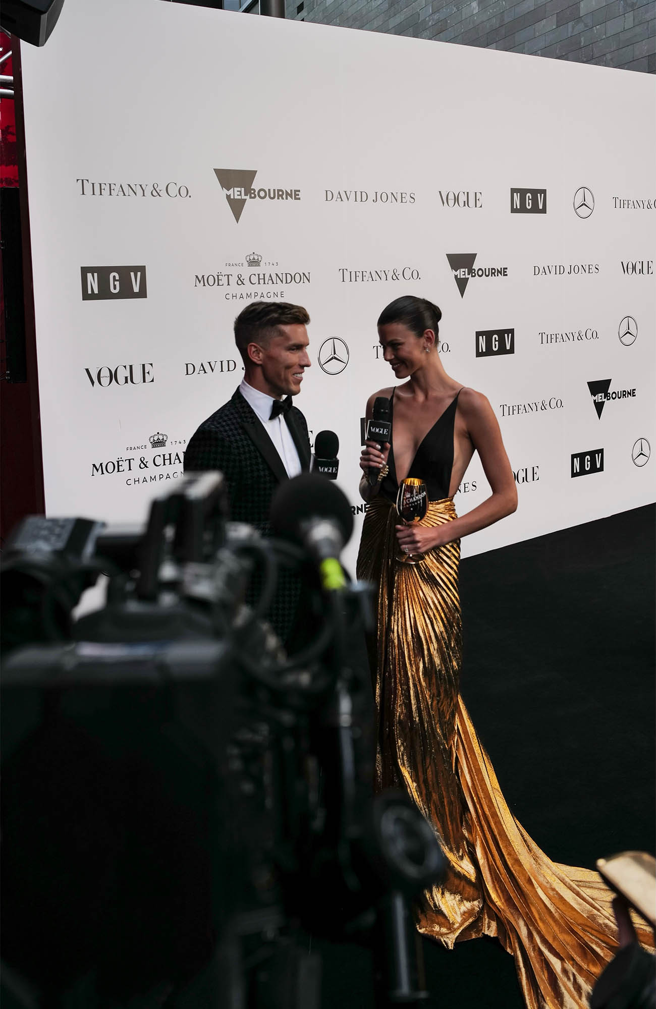 Two people standing in front of a wall with brand logos. A man in a tuxedo interviews a woman in a golden gown; camera in foreground.