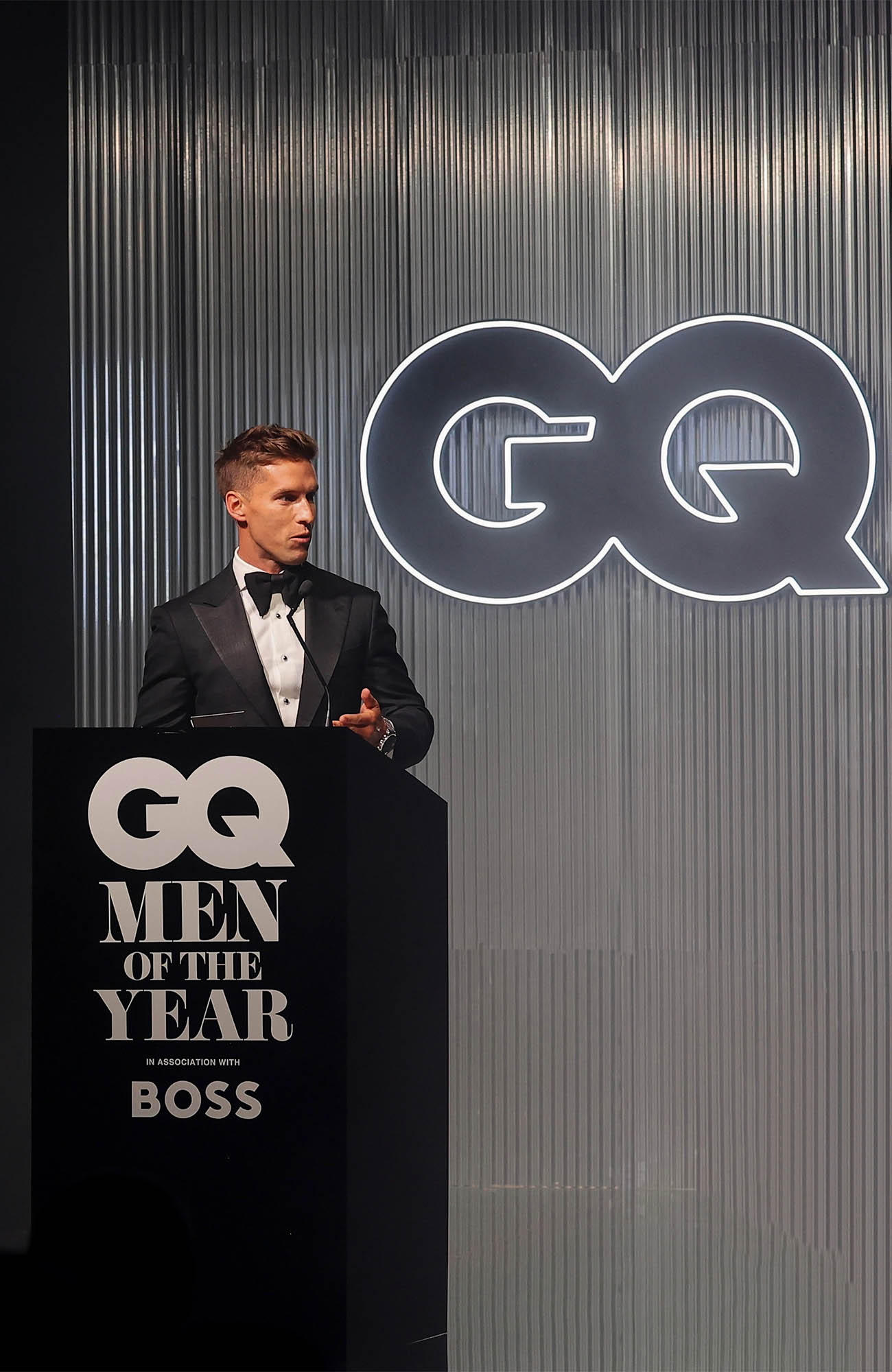 Adult male in a black tuxedo standing at a podium with GQ logos during an indoor event featuring a ribbed silver wall.