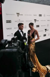 Man in tuxedo and woman in gold dress being interviewed in front of a branded media wall by Matt Suleau