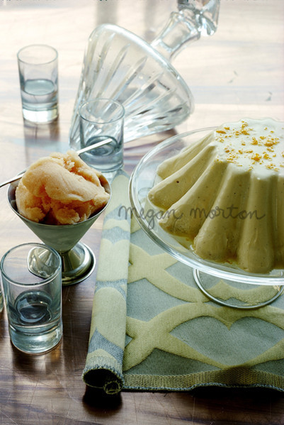 Dessert food photography: A light-yellow molded pudding on a cake stand, sherbet in a silver bowl, and drinking glasses.