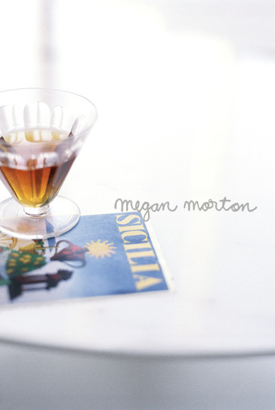 High-angle shot: a cocktail glass with brown liquid and a travel magazine, ‘Sicilia,’ sit on a white round table by a window.