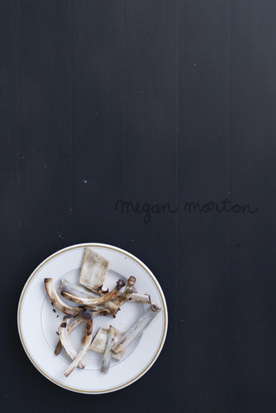 A white plate with bone scraps sits on a dark background with the text 'megan morton' handwritten above the plate, capturing a stark and minimalist composition.