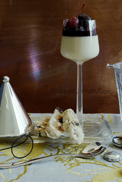 Elegant dessert in stemmed glass with cherries, biscotti, and silver accents on a patterned tablecloth, set against a copper backdrop.
