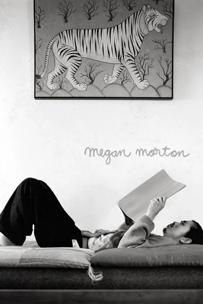 Monochrome interior portrait of a person lying down reading a book beneath a framed tiger painting styled by Megan Morton.