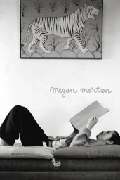 Monochrome interior portrait of a person lying down reading a book beneath a framed tiger painting styled by Megan Morton.