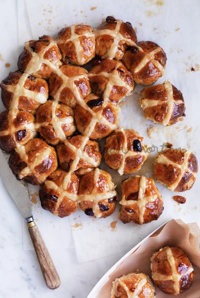 Overhead food styling shot of glazed hot cross buns arranged on white parchment paper with a butter knife and extra buns in a basket.
