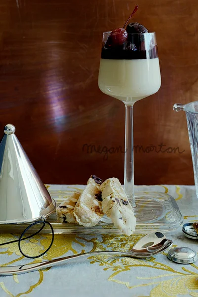Panna cotta in a stemmed glass topped with cherries and biscotti on a patterned tablecloth styled by Megan Morton.