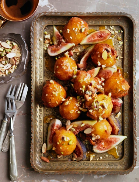 An overhead food styling shot of honey-drizzled fried pastries with fresh fig slices and chopped nuts on a decorated brass tray.