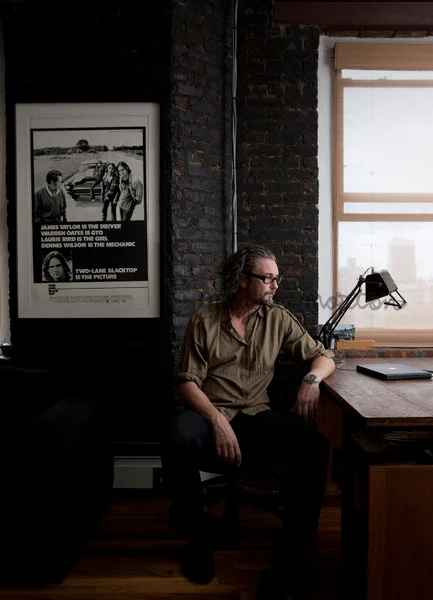 Man in olive shirt and glasses seated at a wooden desk in an interior styled by Megan Morton.