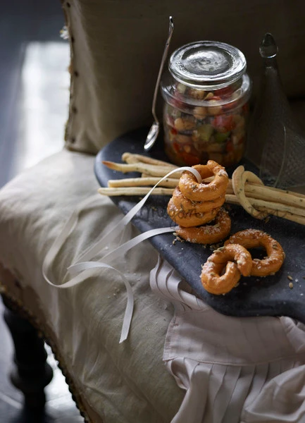 Stacked ring-shaped savory pastries and breadsticks on a dark platter with a glass jar of vegetables, styled by Megan Morton.