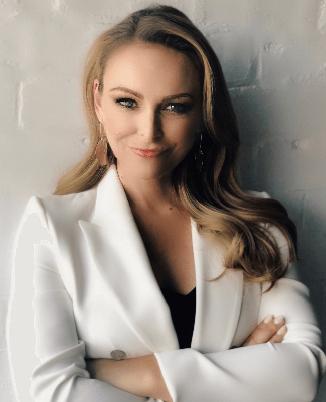 A confident woman with long blonde hair poses indoors in a white blazer. She's standing in front of a textured white brick wall.