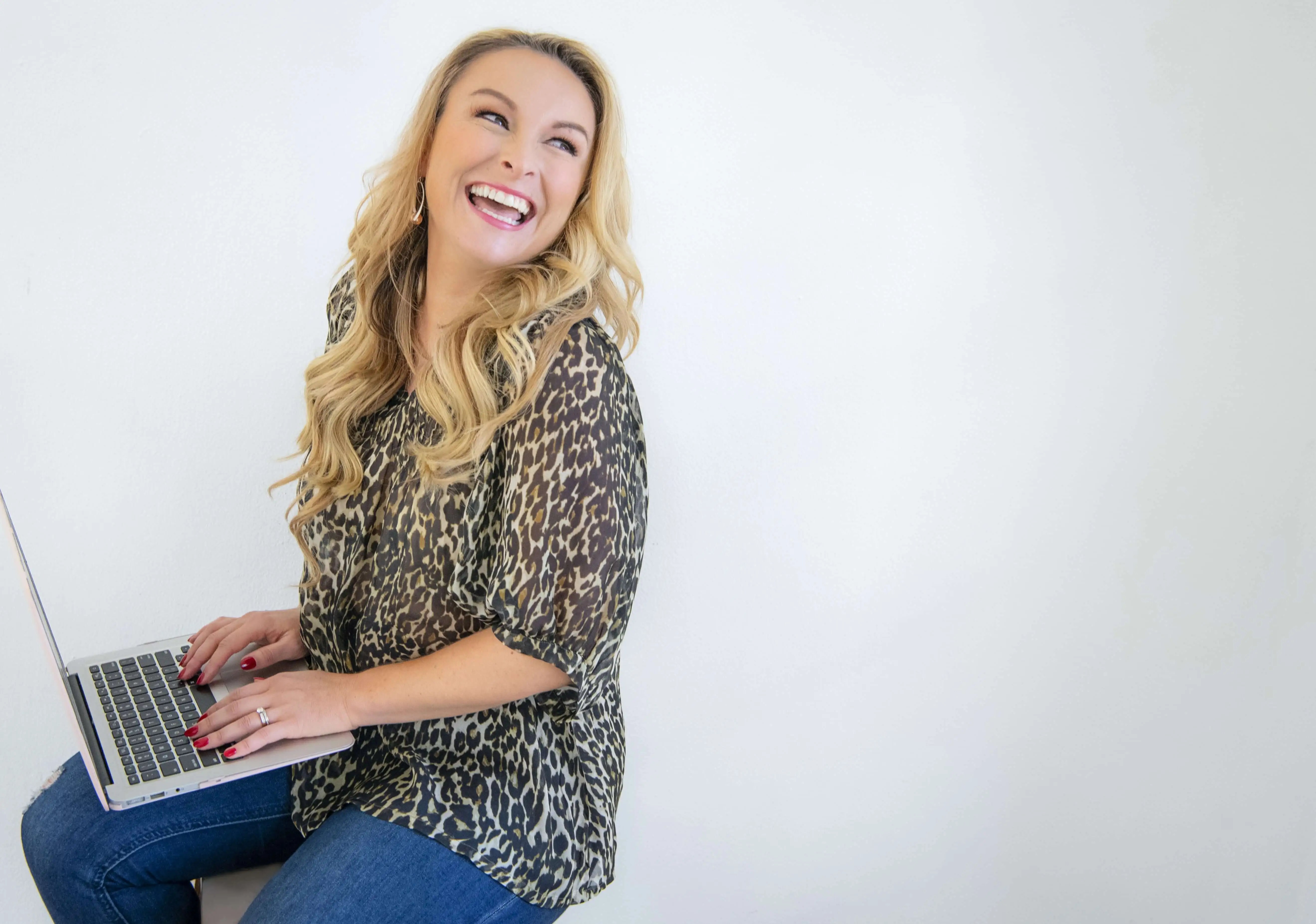 Blond woman sits with a laptop, smiling and looking away, wearing a leopard-print blouse and jeans against a plain white background.