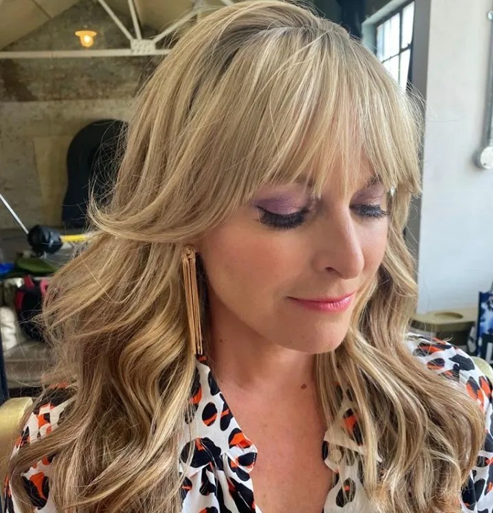 Close-up of a blonde adult female with bangs, purple eye shadow, and statement earrings, wearing a patterned blouse in an indoor environment.