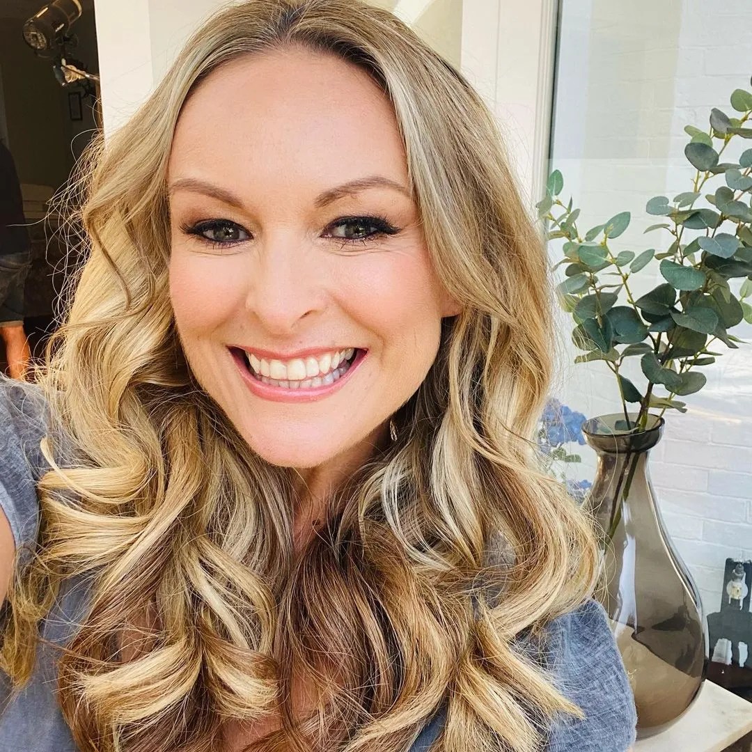 Close-up portrait of a smiling adult woman with long, wavy blonde hair indoors, next to plants in a vase.