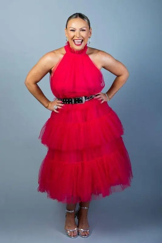 Full-length portrait of a smiling, middle-aged woman wearing a red tulle dress with a black belt against a grey studio background.
