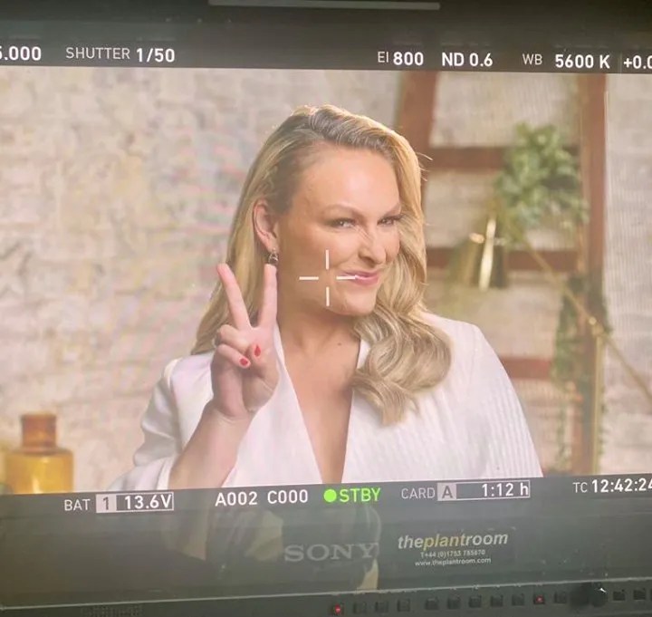 Blonde woman in white blazer gives peace sign with fingers, smiling indoors with plants in the background, taken on camera equipment.