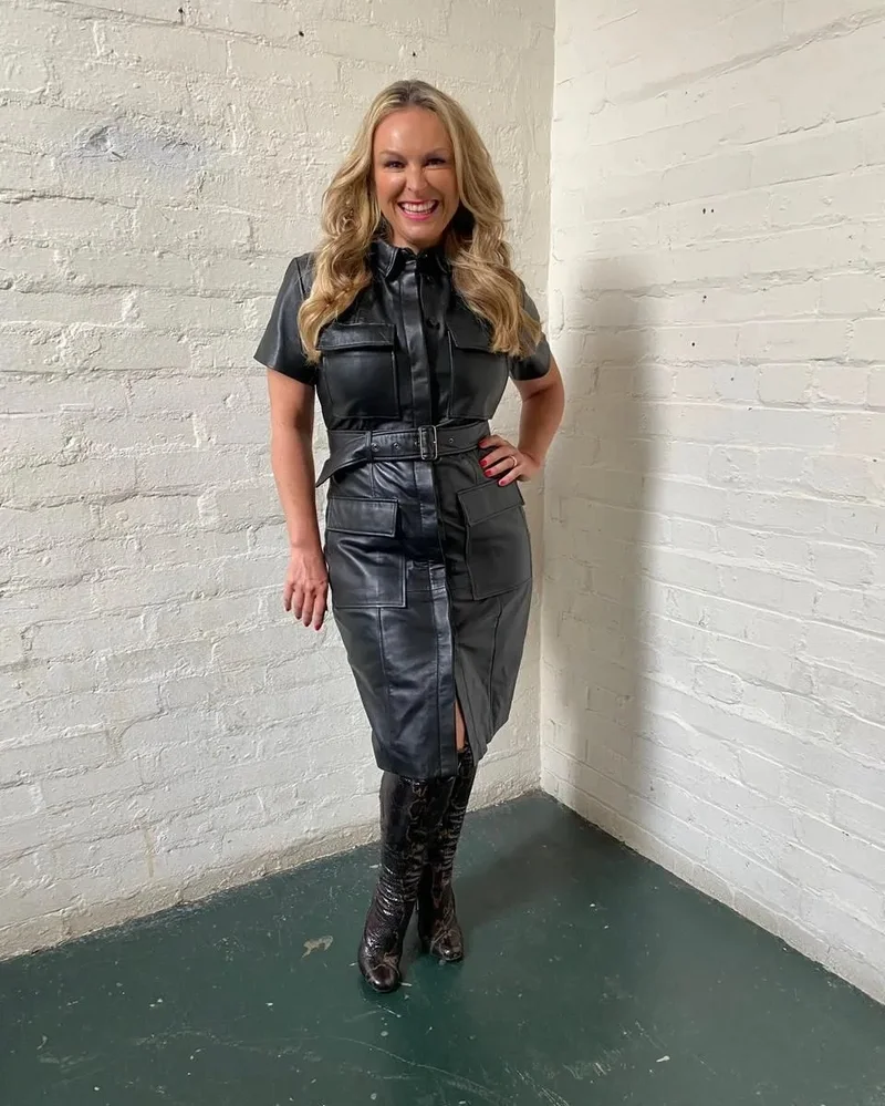 Smiling woman in a black leather shirt dress and tall boots standing against a painted white brick corner in Sydney.