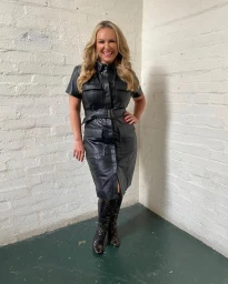 Smiling woman in a black leather shirt dress and tall boots standing against a painted white brick corner in Sydney.