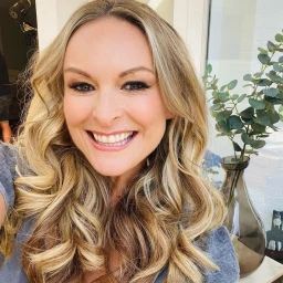 Portrait of smiling blonde woman with long wavy hair wearing a grey top in a brightly lit indoor space