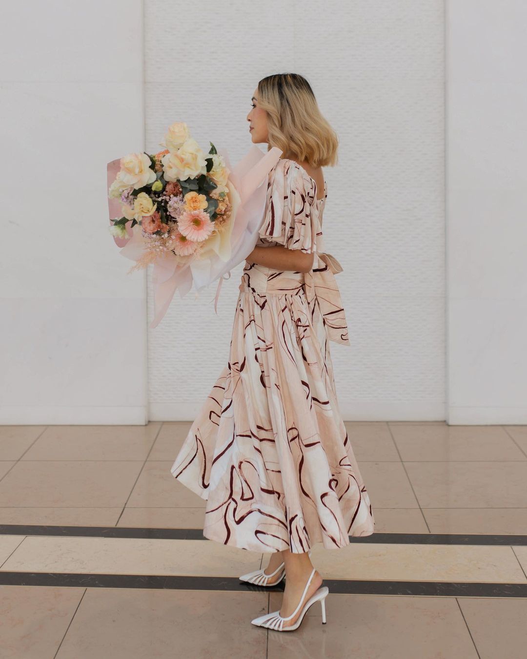 Woman in patterned dress poses indoors holding a bouquet of flowers, wearing high heels, against a white wall with neutral-toned flooring.