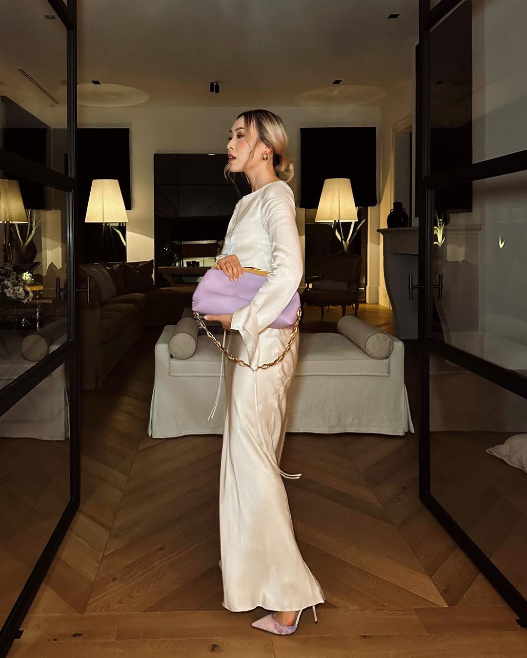 Adult woman poses in long, cream-colored dress with light purple handbag indoors on hardwood floor, near door, and soft lighting.