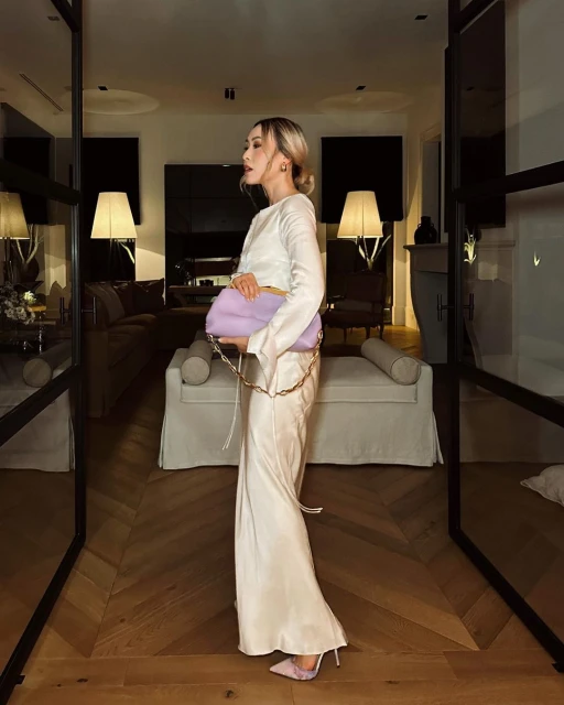 Adult woman poses in long, cream-colored dress with light purple handbag indoors on hardwood floor, near door, and soft lighting.