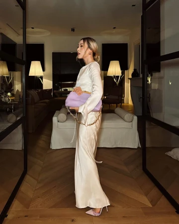 Adult woman poses in long, cream-colored dress with light purple handbag indoors on hardwood floor, near door, and soft lighting.