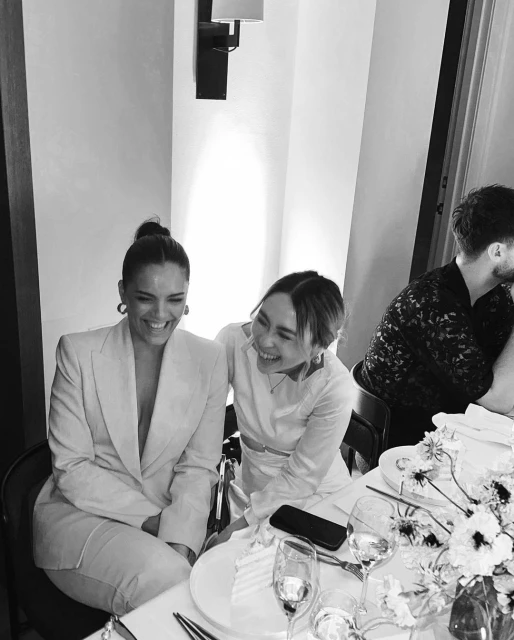 Two women share a laugh at a table set with glassware, plates, and flowers, bathed in soft, neutral lighting, and featuring a third person.