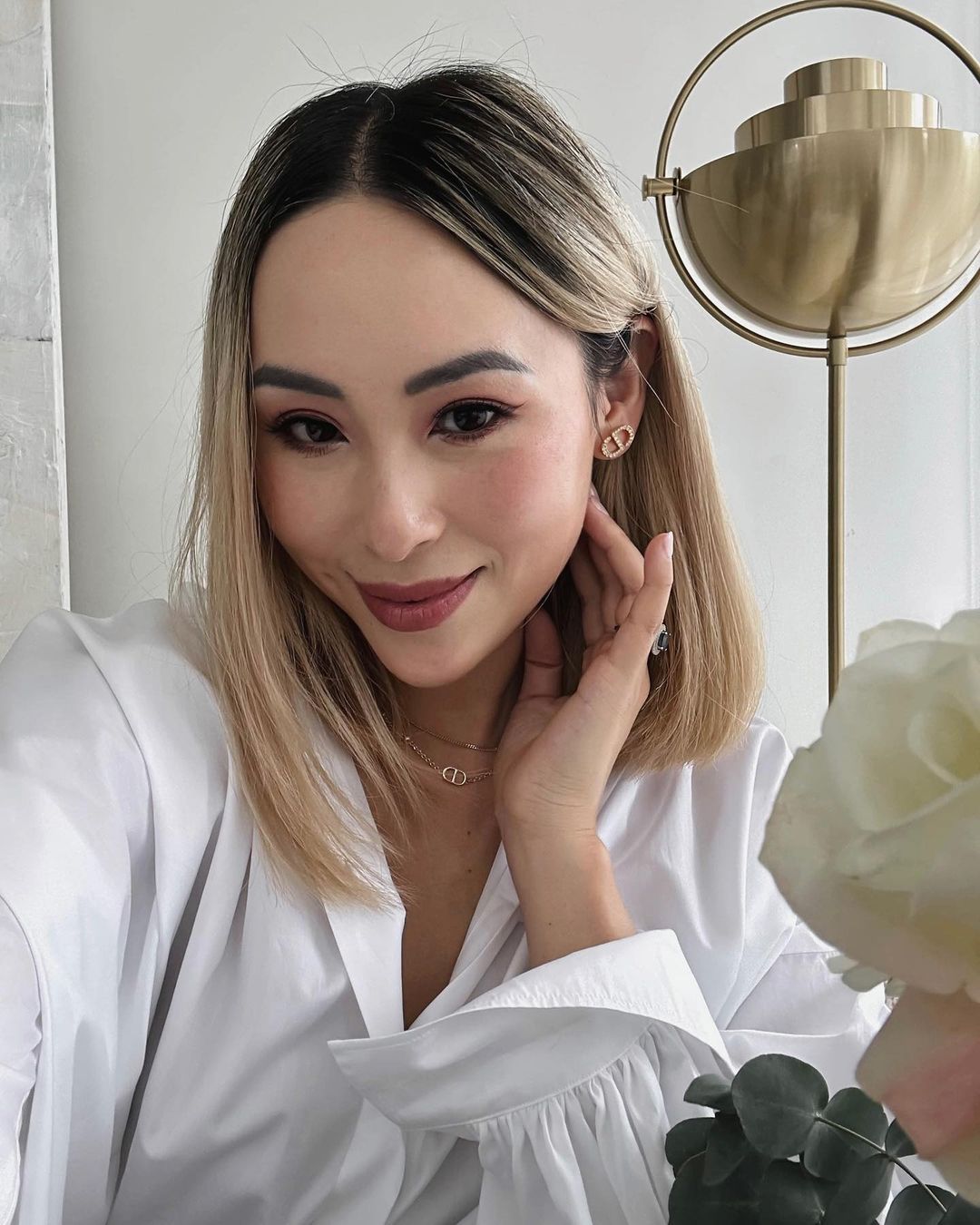 Adult woman in white shirt wearing Dior jewelry, poses in an eye-level, medium shot, with indoor background, soft lighting and flowers.
