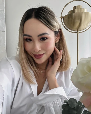 Adult woman in white shirt wearing Dior jewelry, poses in an eye-level, medium shot, with indoor background, soft lighting and flowers.