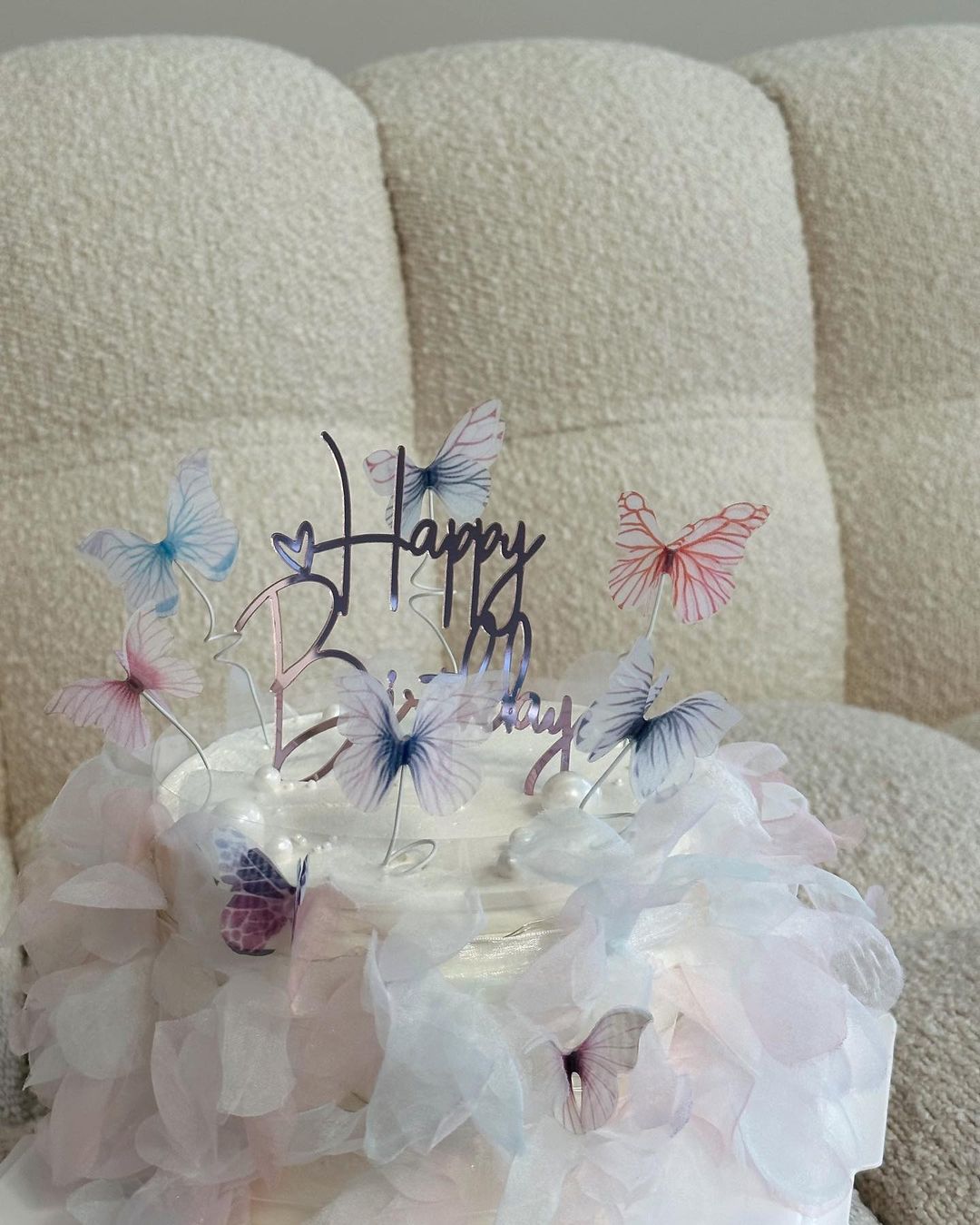 Celebratory cake with white frosting, delicate wafer paper decor, butterfly toppers, and 'Happy Birthday' message on a cream-colored sofa.