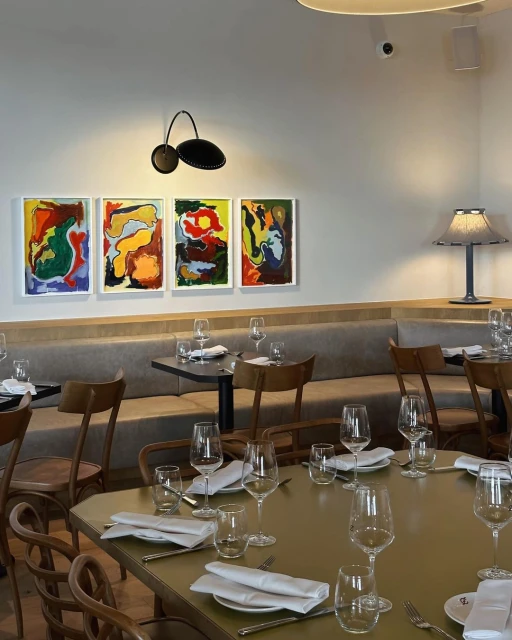 A restaurant interior with art on the wall, set tables with wine glasses and folded white napkins, and booth seating.