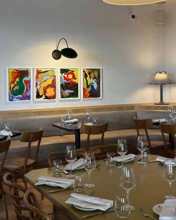 A restaurant interior with art on the wall, set tables with wine glasses and folded white napkins, and booth seating.