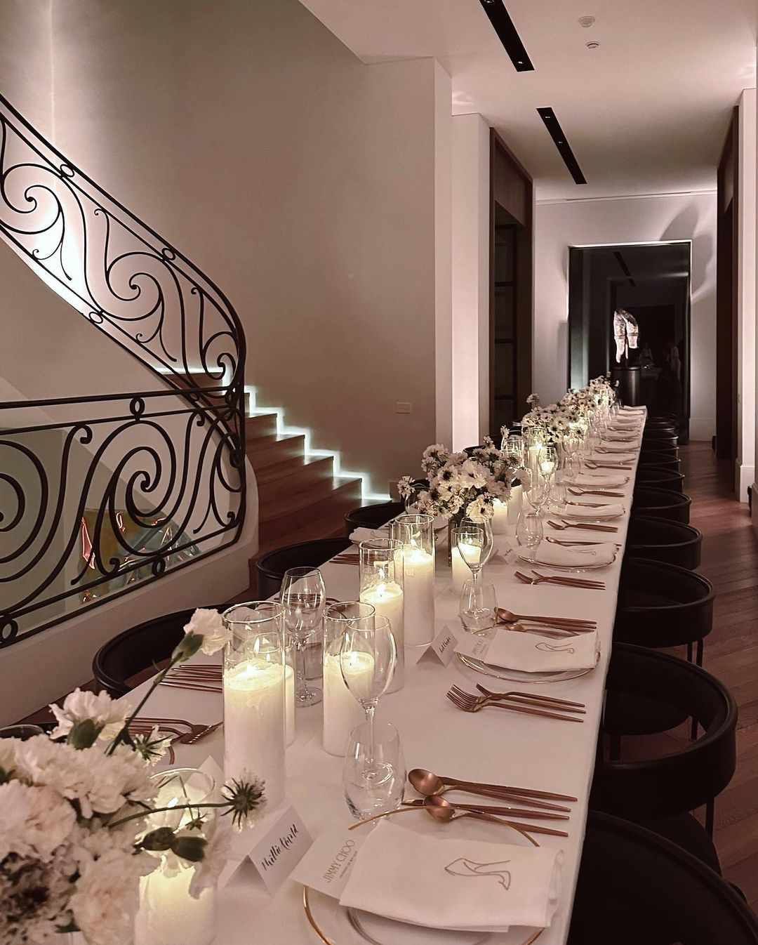 Long formal dining table set with candles, flowers, and place settings, featuring elegant floral arrangements and staircase.