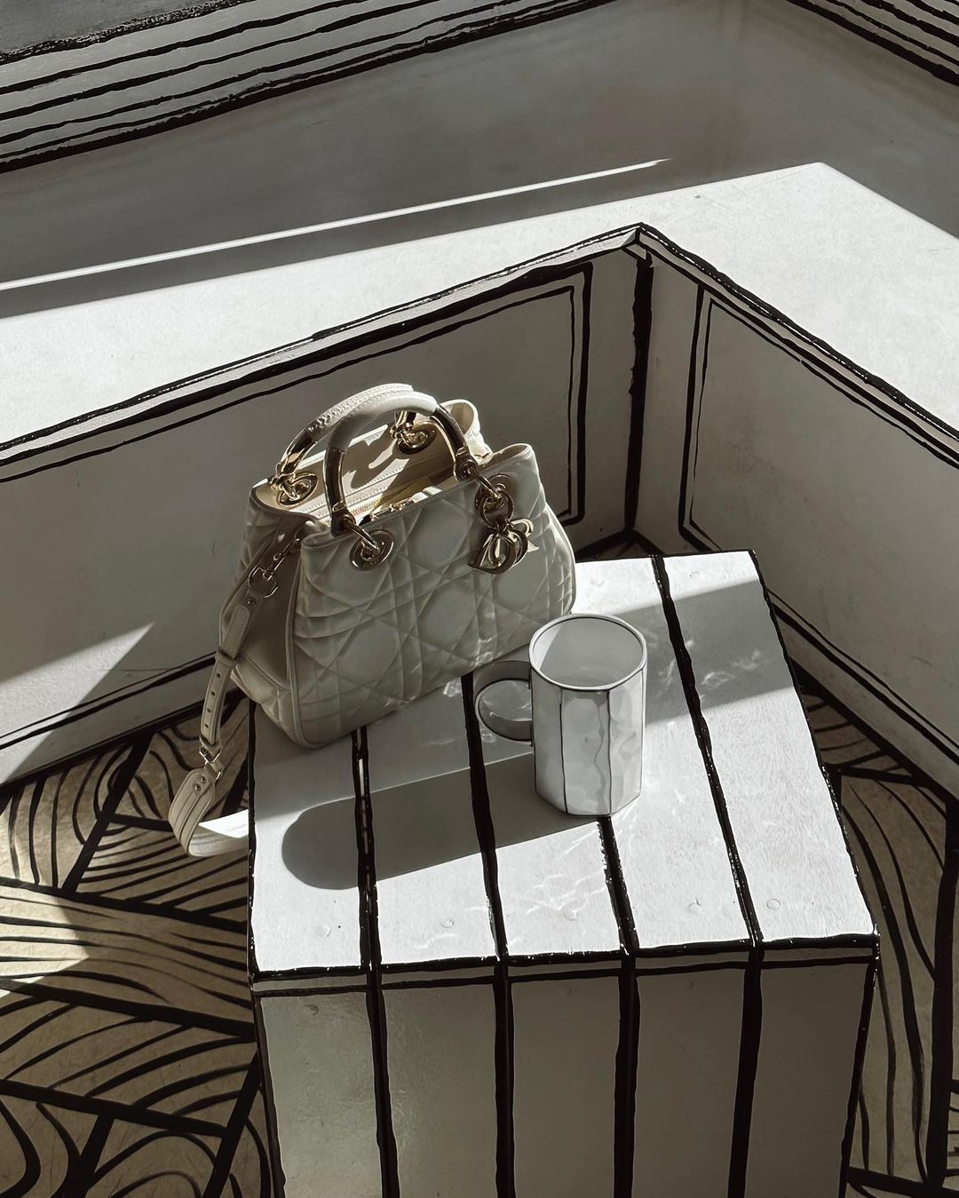 Cream-colored Lady Dior handbag on a striped cube with a white mug, set against a matching striped background.