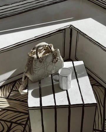 Cream-colored Lady Dior handbag on a striped cube with a white mug, set against a matching striped background.