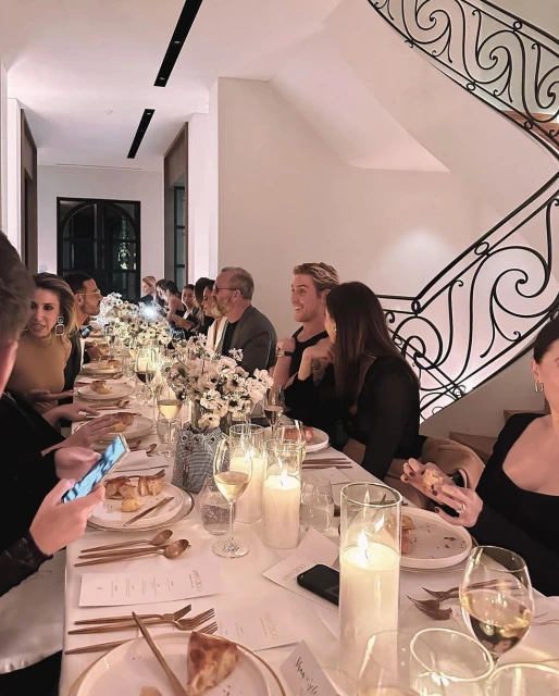 Indoor dinner party scene at long table with several adults eating, drinking, and using phones, illuminated by candles, flowers, and overhead lighting.