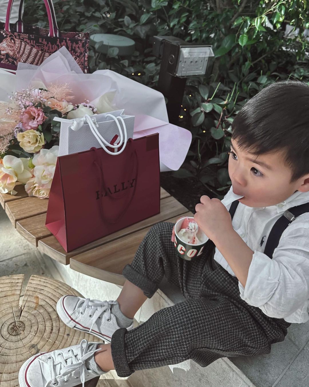 Toddler enjoying ice cream outdoors sitting on bench next to designer bags and a bouquet; dressed in Converse sneakers and rolled trousers.