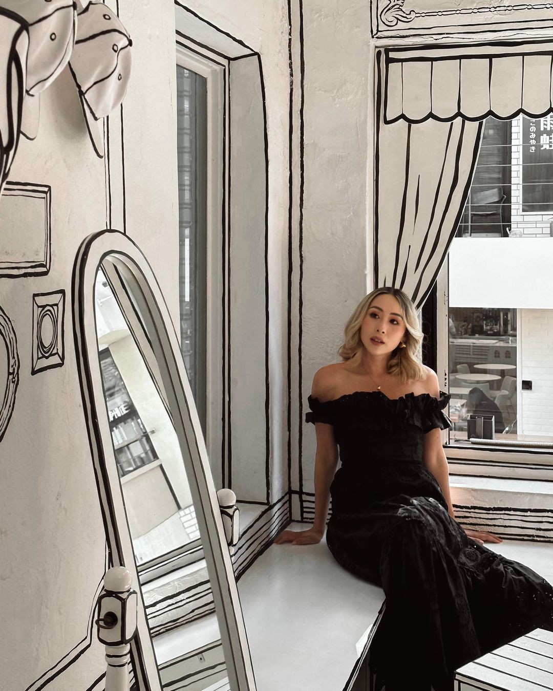 Blonde woman in a black dress poses in a white room with black line art, sitting on a ledge next to a decorative mirror.