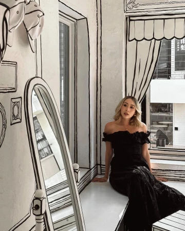 Blonde woman in a black dress poses in a white room with black line art, sitting on a ledge next to a decorative mirror.