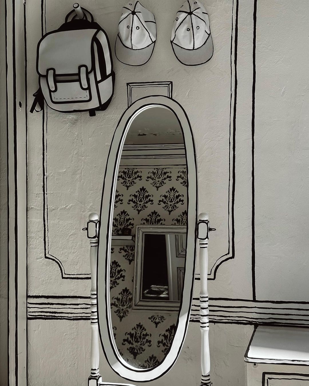 Mirror and wall art featuring a cartoon-style backpack and caps hanging above the standing mirror in a white-and-black interior setting.